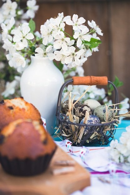 A close-up shot of a DIY Easter centerpiece. It features a wooden crate filled with assorted pastel-colored eggs, small potted tulips, and faux moss. A burlap ribbon is tied around the crate, and a hand-painted wooden sign that reads 