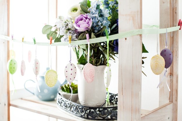 A brightly lit craft table with various DIY Easter decoration supplies, including pastel-colored paints, paintbrushes, colorful yarn, ribbons, and unfinished wooden cutouts of bunnies and eggs. Hands are seen decorating Easter eggs with intricate patterns and designs, showcasing a creative and hands-on Easter activity.